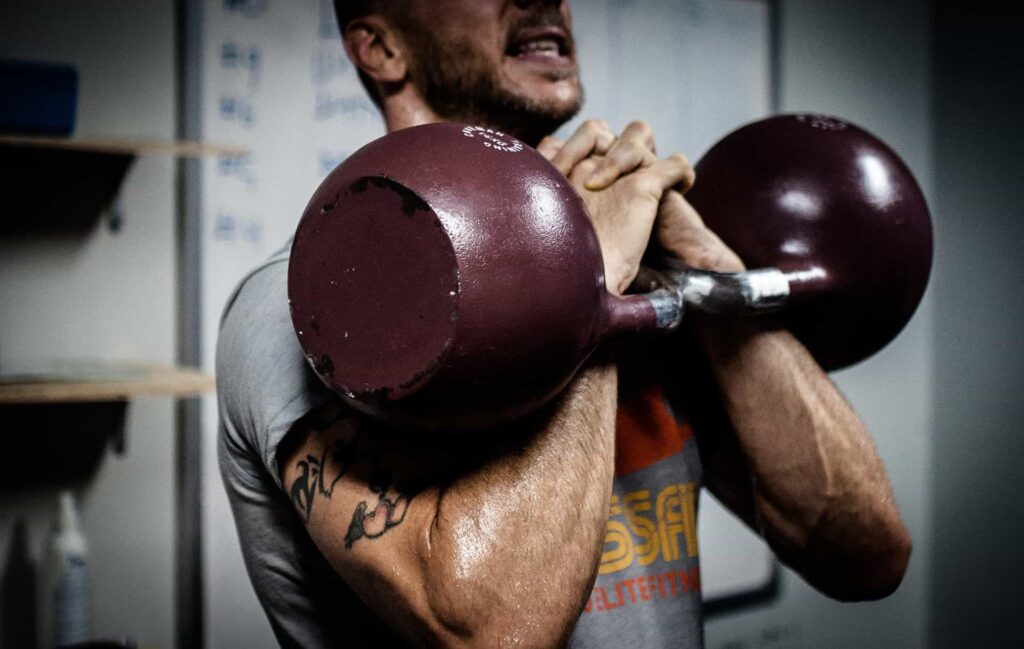 A man standing and lifting kettlebells in front of him to build muscle mass.