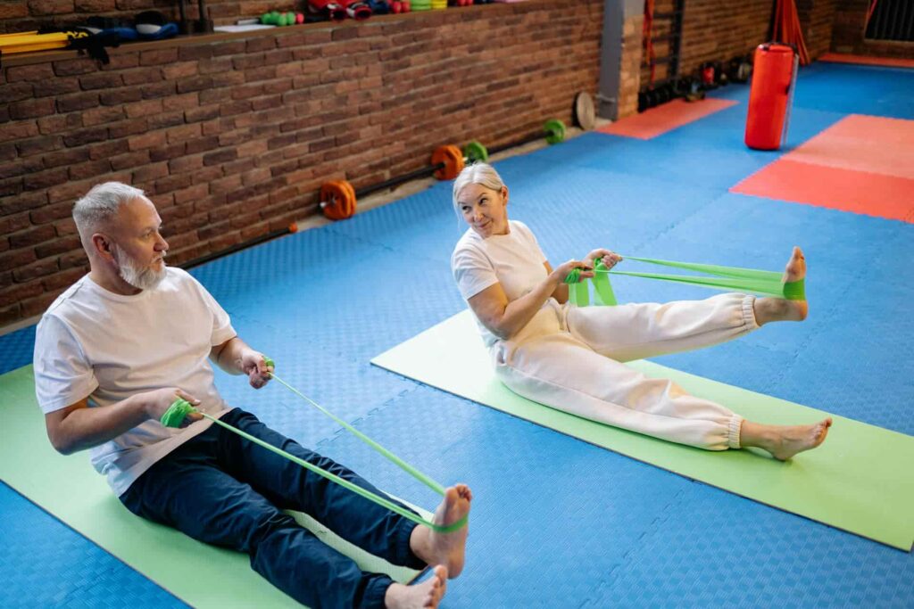 Elderly couple performing resistance band exercises on mats as part of balance training for seniors in a gym setting.