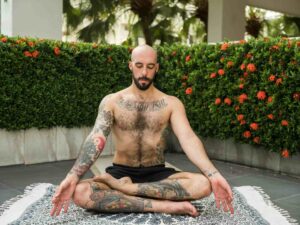 A tattooed man meditating outdoors in a cross-legged position, practicing mindfulness-based stress reduction on a patterned mat surrounded by greenery and red flowers.