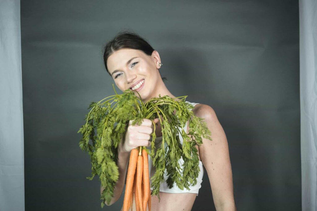 A woman holding carrots and smiling, showcasing foods that are great for your skin.